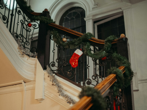 A staircase decorated for the holidays, featuring a garland of greenery wrapped around the railing with red and gold ornaments. A festive red Christmas stocking with white trim is hung on the railing. The backdrop includes white walls and arched windows, creating a cosy and elegant holiday atmosphere.