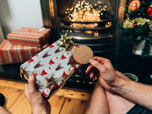 A pair of hands holding a gift wrapped in festive paper featuring Santa Claus and reindeer illustrations, topped with a gold bow and a tag labeled 'Secret Santa.' The background includes wrapped presents, a decorated Christmas tree, and a lit fireplace adorned with warm fairy lights, evoking a cosy holiday scene