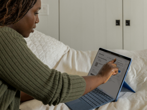 A woman with dark braided hair and a green sweater is sitting on a bed with white bedding. She is using a stylus or finger to interact with a blue tablet screen displaying an online shopping website. The setting is calm, with a white wardrobe in the background.