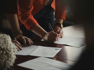 A close-up of two people at a table signing documents. One person is wearing an orange blazer and holding a pen, poised to sign a piece of paper. The other individual, with a watch on their wrist, has their hand resting on the table. The table is covered with documents, and a soft focus on white flowers is visible in the foreground.